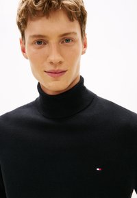 Jonge man met kort krullend haar die een zwarte coltrui draagt met een klein rood, wit en blauw logo op een witte achtergrond.