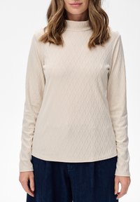 Vrouw in een beige, gebreide top met lange mouwen en een hoge hals, en een donkerblauwe broek, staand tegen een effen witte achtergrond.