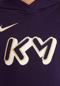 Sudadera morada con un gran logo blanco y dorado que presenta las letras "KM". El material parece suave y texturizado, con capucha ajustable.