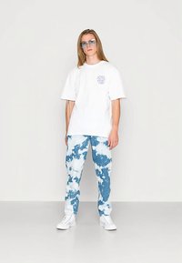 Balti medvilniniai marškinėliai su mėlynu logotipu ant krūtinės, derinami su mėlynais džinsais su tie-dye efektu ir baltais sportbačiais, stovint neutralioje aplinkoje.