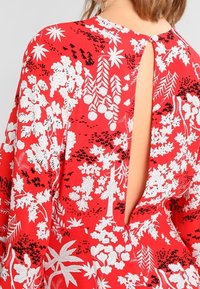 Robe rouge à motifs floraux blancs, avec une découpe profonde dans le dos et une fermeture à boutons au niveau du cou. Fabriquée en tissu léger.
