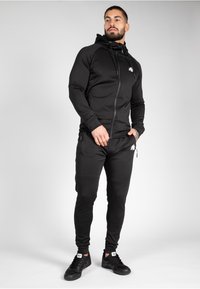 Tuta sportiva nera con felpa con zip e pantaloni aderenti. Tessuto morbido ed elastico; include il logo sul petto e tasche laterali.
