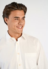 Jonge man met donker haar die glimlacht, gekleed in een witte overdent shirt met een zak, die iets naar rechts kijkt tegen een effen achtergrond.