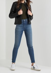 Černá kožená bunda, žebrovaný zelený svetr, modré džíny s vysokým pasem a úzkým střihem a bílé tenisky s černými detaily. Model stojí před jednoduchým pozadím.