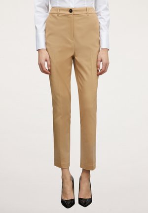 Vrouw draagt beige op maat gemaakte enkel-lange broek, wit overhemd met lange mouwen en zwarte puntige hoge hakken, staand tegen een effen achtergrond.