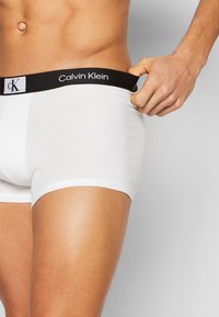 Baltos medvilninės bokserių kelnaitės su juoda elastine juosta. Juosta puošia „Calvin Klein“ logotipas. Lygus paviršius, prigludęs dizainas ir iki vidurinės šlaunies ilgio.