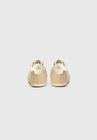 Beige sneakers with a smooth suede finish, white accents, three stripes, and a padded heel. Minimalist design with a low profile.