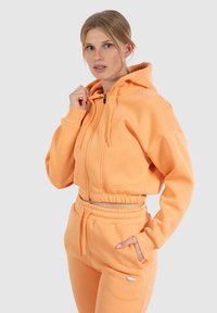 Jeune femme portant un hoodie court pêche et un pantalon de survêtement assorti, les mains dans les poches, devant un fond gris clair uni.