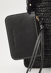 Sort vævet håndtaske med en struktureret krop. Har et glat sort lædermærke, fastgjort med en tynd rem med frynser. Guld lynlåsdetalje.