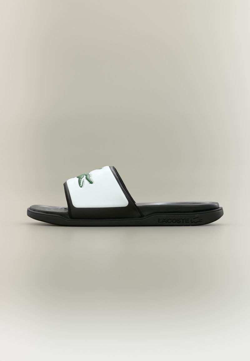 Svart och vit slide sandal med en slät gummimaterial, med ett brett band som visar en grön krokodil-logotyp och en texturerad sula.