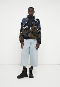 Kurtka polarowa w niebiesko-zielono-brązowy wzór górski, z zamkiem z przodu i wysokim kołnierzem, zestawiona z luźnymi jasnoniebieskimi dżinsami i czarnymi butami.