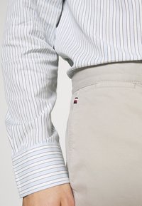 Svetle béžové kalhoty s hladkým povrchem, s malým červeným, bílým a modrým logem na zadní kapse. Je vidět pruhovaný modro-bílý rukáv košile.