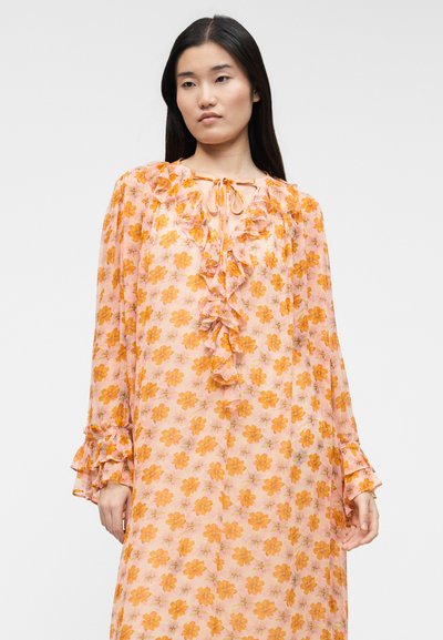 Γυναίκα με μακριά σκουρόχρωμα μαλλιά που φοράει ροζ διαφανές φόρεμα με πορτοκαλί ανθοπωλεία, γιακά με βολάν και φουσκωτά μανίκια, στέκεται μπροστά σε απλό φόντο.