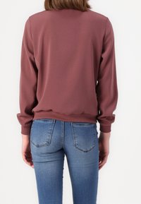 Bordeauxrode sweatshirt met lange mouwen en geribde manchetten; gladde textuur. Draagt over blauwe jeans, met achterzakken en een aansluitende snit.