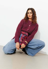 Pull en Tricot bourgogne avec texture côtelée et trois boutons, associé à un jean large bleu clair et une chemise à carreaux rouge et bleu nouée à la taille.