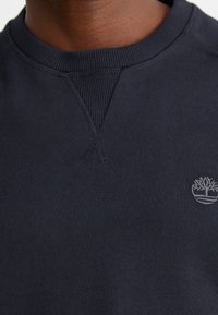Gros plan d'une personne portant un sweat-shirt bleu marine foncé avec un col côtelé et un petit logo arbre gris clair sur la poitrine.