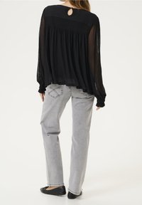Blouse noire transparente à coupe ample, dos smocké, détail œillet au col, associée à un jean gris clair à jambes droites et des ballerines noires.
