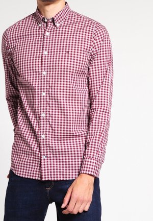 Homme portant une chemise ajustée à manches longues à carreaux rouges et blancs avec un jean bleu sur un fond uni.