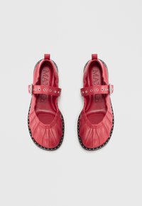 Rote Lederschuhe mit gerafftem Design, verstellbarem Riemen und dekorativen Nieten entlang der schwarzen Sohle. Abgerundete Zehenform.