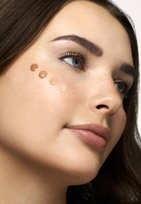 Muestras de base líquida en varios tonos beige en la mejilla; textura de piel suave, reflejos de luz que realzan un aspecto natural.