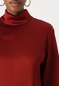 Top rojo de satén con cuello alto y una textura suave y brillante; presenta un ajuste relajado y costuras detalladas en los hombros.