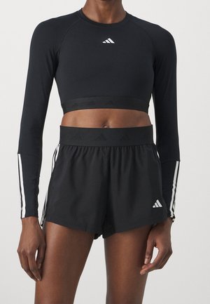 Atletisk person iført sort langærmet crop top og sorte løbeshorts med hvide Adidas-logoer og striber på ærmer og shorts.