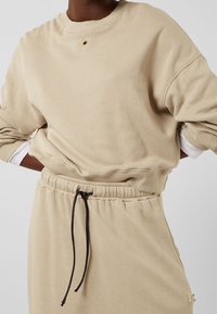 Sweat-shirt beige avec un col rond, des épaules tombantes et des poignets côtelés, présentant un détail œillet central et une taille élastiquée avec un cordon de serrage.