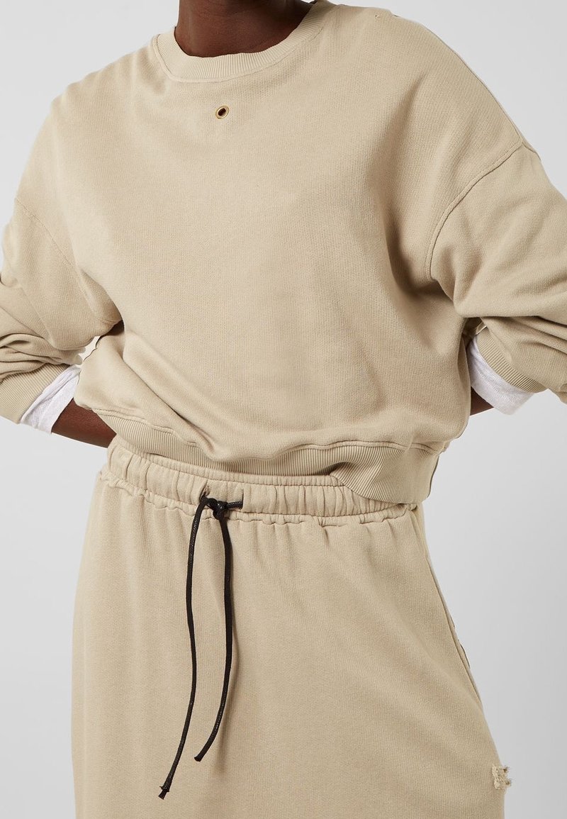 Sweat-shirt beige avec un col rond, des épaules tombantes et des poignets côtelés, présentant un détail œillet central et une taille élastiquée avec un cordon de serrage.