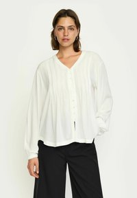 Blusa bianca a maniche lunghe con scollatura a V, pieghe verticali e chiusura con bottoni. Presenta polsini elasticizzati e una forma ampia e fluida. Abbinata a pantaloni neri.