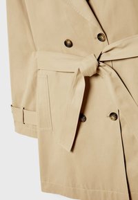 Trench beige avec boutons croisés, poche latérale, taille ceinturée nouée en nœud et détail de bride sur le poignet de la manche.