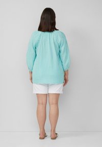 Himmelbluse mit weißen Streifen, langen Ärmeln und gerafftem Ausschnitt, kombiniert mit weißen, hochgekrempelten Shorts, von hinten betrachtet.