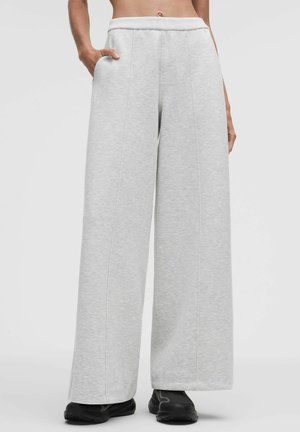 Pantaloni da ginnastica grigio chiaro a gamba larga con cuciture verticali, vita elastica, tasche laterali, indossati con scarpe sportive nere da una persona.