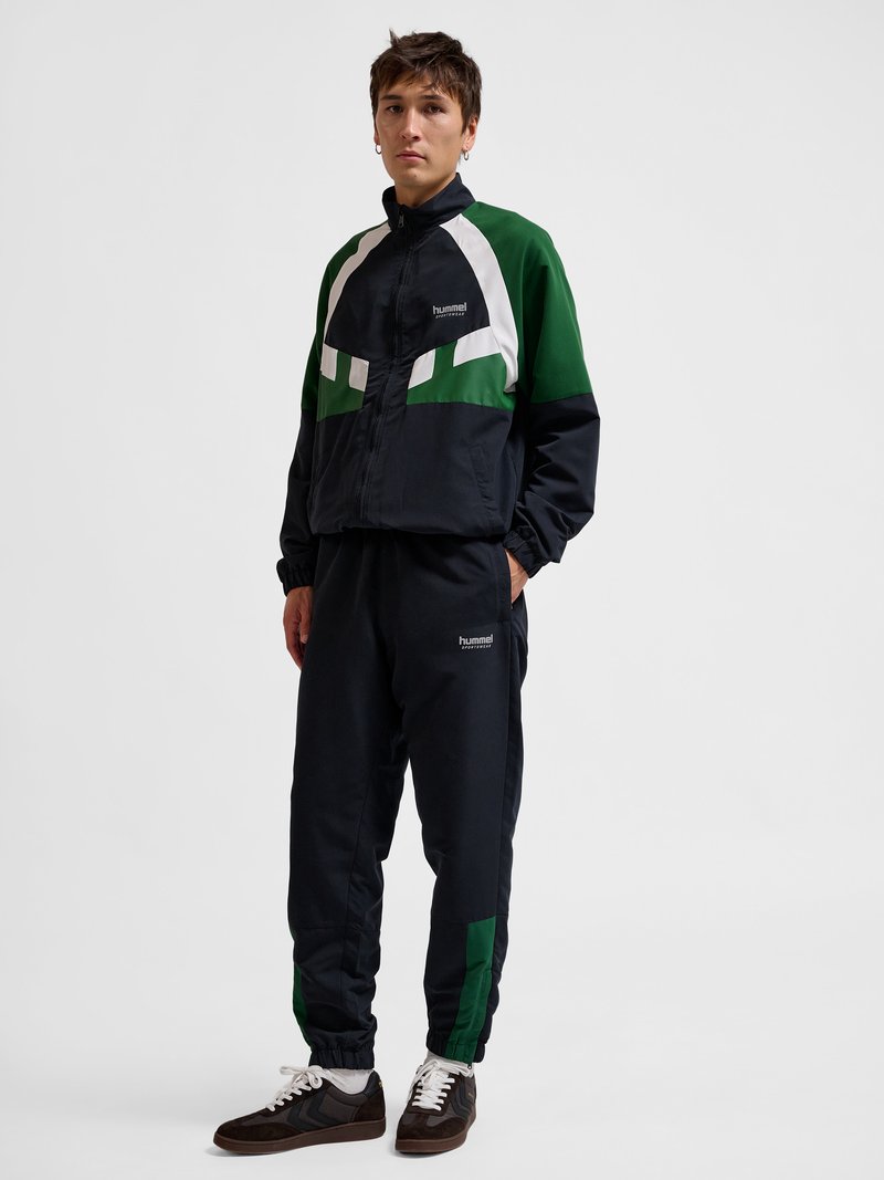 Młody mężczyzna w czarno-zielono-białym dresie Hummel z kurtką na zamek i spodniach dresowych, stojący z rękami w kieszeniach.