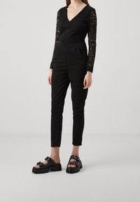 Svart spetsbodysuit med V-ringning, långa ärmar och åtsittande svarta byxor. Svarta plattformssandaler med spännen och strukturerad sula.