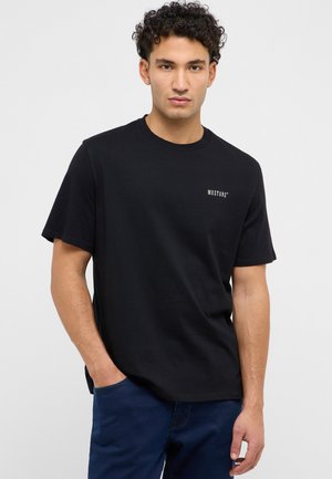 Młody mężczyzna z kręconymi włosami w czarnej koszulce Mustang i niebieskich spodniach, stojący z jedną ręką w kieszeni na jednolitym tle.