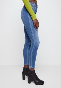 Femme portant un jean skinny bleu clair, des bottines à talons noires et un crop top vert à manches longues, debout devant un fond blanc.