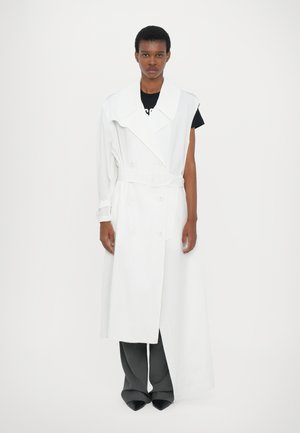 Manteau trench oversized blanc avec un large col, une ceinture et des boutons à double boutonnage, superposé sur une chemise noire et un pantalon gris.