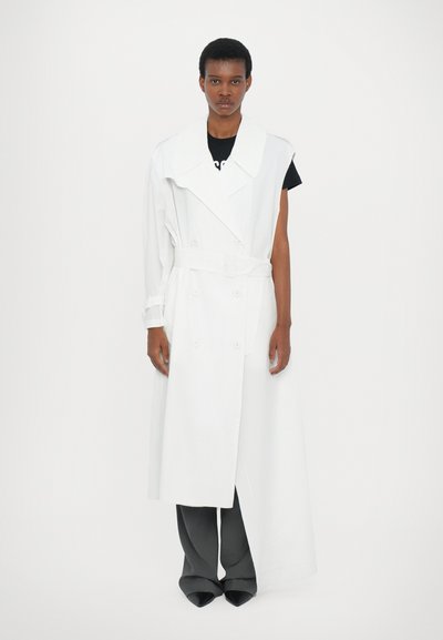 Manteau trench oversized blanc avec un large col, une ceinture et des boutons à double boutonnage, superposé sur une chemise noire et un pantalon gris.