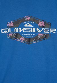 Sinine T-särk kuuekülgse Quiksilveri logoga, millel on tumevärviline lillemuster ja tekst "TÕELINE BOARDRIDINGU ETTEVÕTE."