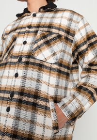 Flanellskjorta-jacka med ett svart, vitt och orange rutmönster, knappstängning, bröstficka och mjuk textur med en svart krage.