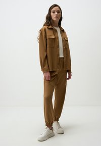 Veste en polaire marron avec deux poches poitrine, assortie à un pantalon ajusté. Le mannequin porte une chemise claire et des baskets blanches. Textures simples et lisses.