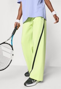 Παντελόνια φαρδιά σε νέον πράσινο με μαύρες λωρίδες στο πλάι, συνδυασμένα με ανοιχτό μωβ πουκάμισο και μαύρα αθλητικά παπούτσια, κρατώντας ρακέτα τένις.