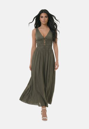 Donna che cammina in avanti indossando un abito lungo senza maniche verde oliva con gonna plissettata e tacchi bianchi, su sfondo chiaro uniforme.