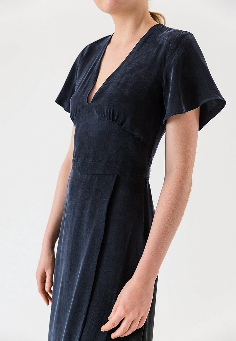 Námořnické modré šaty s hlubokým véčkovým výstřihem, krátkými rukávy a vypasovaným tělem. Vyrobené z hladké látky s jemným leskem. Detail s sklady na sukni.