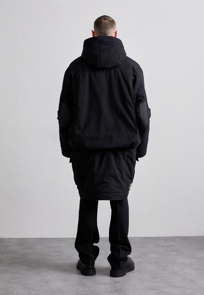 Schwarze Kapuzenjacke mit langem Rücken, aus strukturiertem Stoff hergestellt. Verfügt über Aufnäher an den Schultern und ein kleines grünes Logo-Detail.