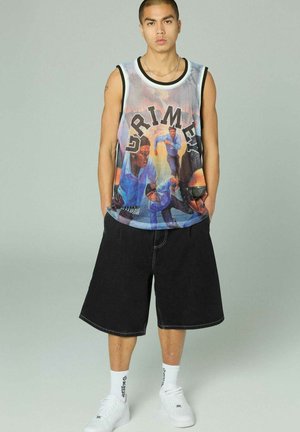 Junger Mann trägt ein ärmelloses Basketball-Trikot mit Grafik, schwarze weite Shorts, weiße Socken und weiße Turnschuhe vor einem hellgrauen Hintergrund.