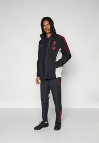 Črno in kremasto športno jakno z rdečimi poudarki, z logotipom Liverpoola. Kombinirana z ujemajočimi se temnimi hlačami in črnimi športnimi čevlji.