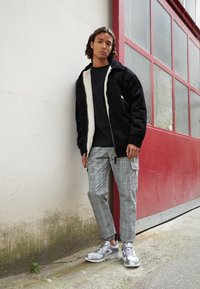 Schwarze Cordjacke mit Fleece-Innenfutter, schwarzes T-Shirt, karierte Cargo-Hosen und silberne Sneakers. Steht an einer strukturierten Wand.