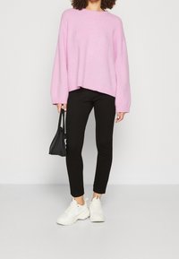 Pink oversized sweater with ribbed hem, paired with black fitted pants and white sneakers. Black handbag held in hand.