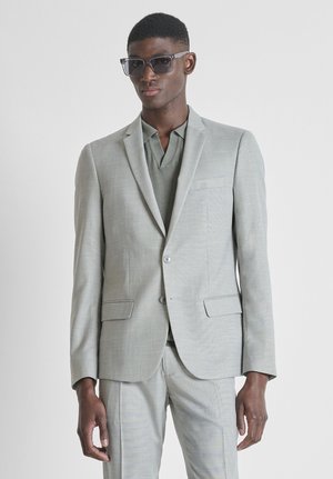 Man in een lichtgrijs pak en een groen poloshirt, met een donkere zonnebril, staand tegen een effen lichte achtergrond.